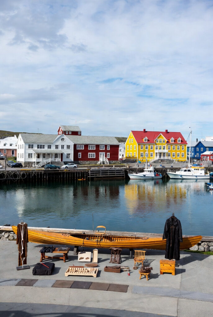 Exterior of the Greenland National Museum in Nuuk, with traditional wooden buildings and colorful colonial architecture beside the harbor. In the foreground, a display of Inuit artifacts (e.g., kayak, sealskin clothing) under soft summer light. Cultural and historical atmosphere.