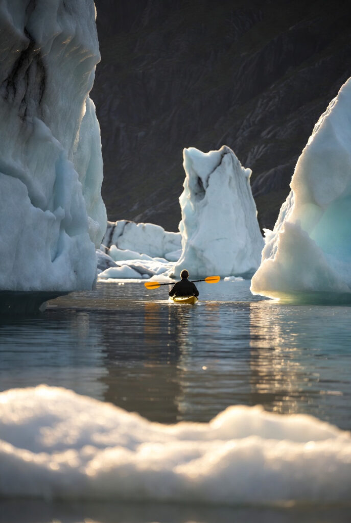  A person in a bright yellow kayak gliding silently between sculpted icebergs in a narrow fjord. Sunlight reflects off the water and ice; distant cliffs rise steeply. Calm, meditative mood, shallow depth of field, documentary-style realism